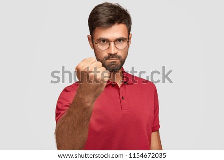 Serious angry male shows fist, ready for fight or challenge, has stern expression, wears casual red t shirt, poses against white background. Aggressive young man gestures indoor. Body language concept