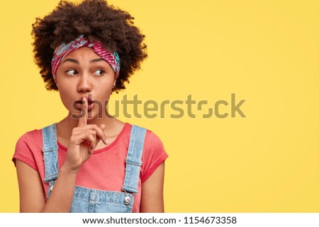 African American female with secret expression, makes silence sign, keeps fore finger on lips, looks aside, poses over yellow background with free space for your promotional content. Hush, be quiet