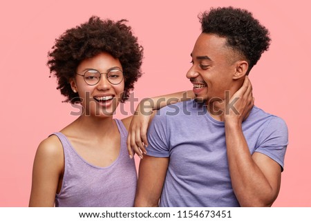 Photo of joyful dark skinned female and male companions have joy together, dressed casually, smile positively, stand against pink background. Happy African American woman leans at shoulder of man Royalty-Free Stock Photo #1154673451