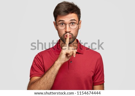 Keep voice down. Attractive surprised bearded male makes shush gesture, demnads shut up, wears casual bright red t shirt, poses against white background. People, hush, conspiracy, secret concept