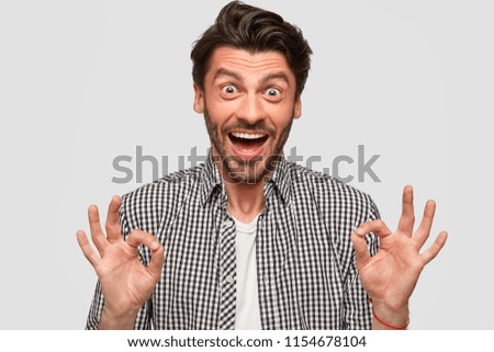 Photo of stylish bearded young male has trendy haircut, makes okay gesture with both hands, dressed in checkered shirt, stares with eyes, isolated over white background. Body language concept