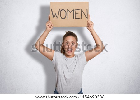 Happy young European lovely feminist dressed in casual grey t shirt, holds plate with inscription Women above head, demonstrates females power, isolated over white background. Feminism concept