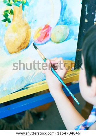 Cute, serious and focused, seven years old boy in blue shirt drawing on canvas standing on the easel. Concept of early childhood education, painting, talent.