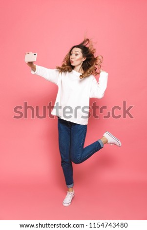 Full length portrait of an excited young woman taking selfie with outstretched hand while jumping isolated over pink background