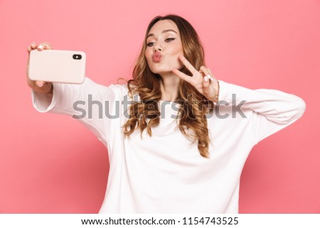 Portrait of a happy young woman taking selfie with mobile phone isolated over pink background, showing peace