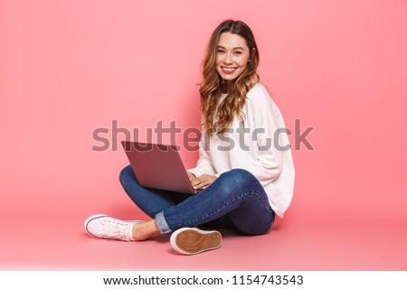 Portrait of a smiling young woman sitting with legs crossed, using laptop computer isolated over pink background