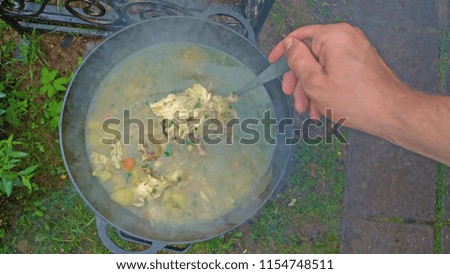 Delicious chicken soup is cooked on an open fire in a black cast-iron pot. A man's hand tries a soup with a ladle. Street food.