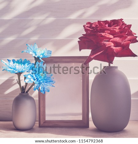 A large red rose is in a gray vase. Nearby is a bouquet of blue chrysanthemums in a vase. Between the vases is an empty wooden frame. White wooden background.