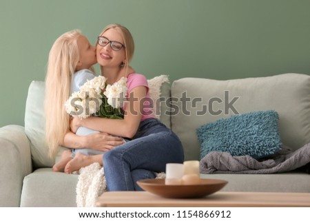 Cute little girl and her mother with beautiful flowers sitting on sofa at home