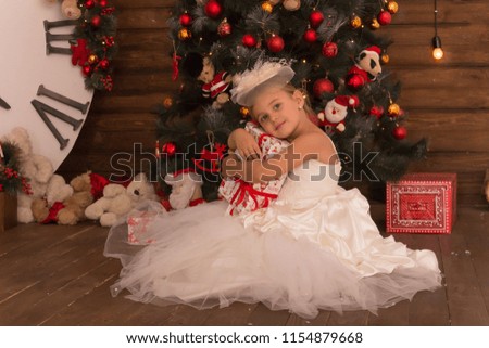 Cute little girl in a white nice dress with a present. Christmas tree, gifts and packs on background. Festive interior. Family holidays