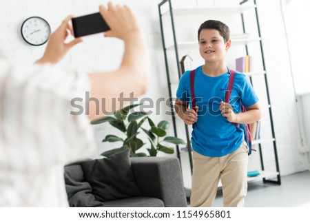 partial view of mother taking picture of son with backpack at home, back to school concept