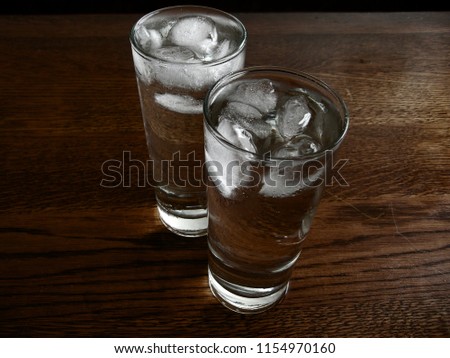 2 water drinks standing on dark brown wood.  Both drinks are cold and have ice. Both are transparant and the glasses are long. Both glasses are full of water. There is little light in this picture.