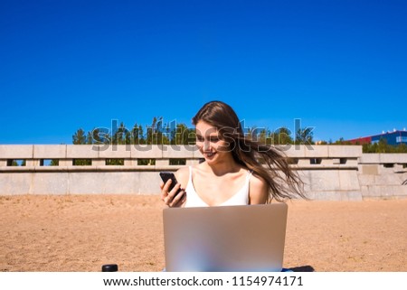 Happy smiling female copywriter watching funny video on mobile phone during distance work on laptop computer, sitting on the beach in sunny summer day against blue sky background with copy space 