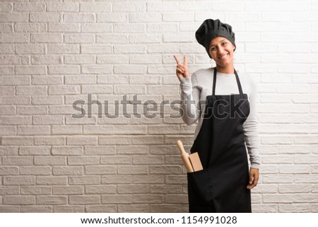 Young baker indian woman against a bricks wall fun and happy, positive and natural, makes a gesture of victory, peace concept