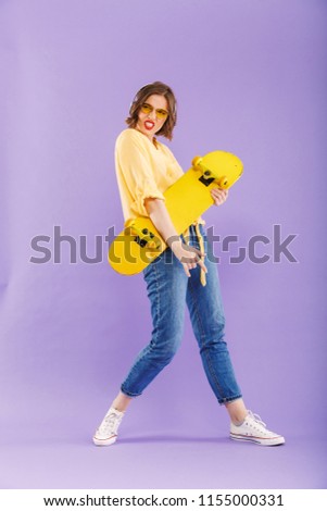 Full length portrait of a cheerful young girl in headphones holding skateboard,standing isolated over violet background
