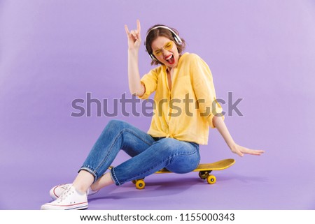 Portrait of an excited young girl in headphones sitting on skateboard isolated over violet background