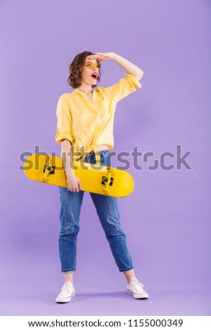 Full length portrait of a cheerful young girl holding skateboard,standing isolated over violet background, looking far away