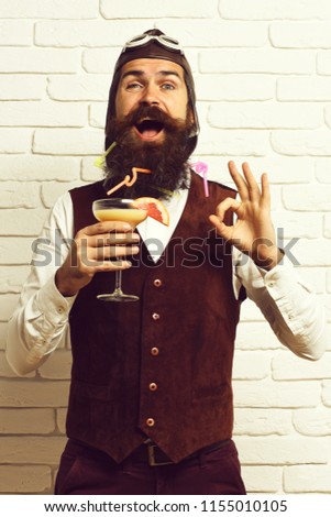 handsome bearded aviator man with long beard and mustache on happy face holding glass of alcoholic cocktail in vintage suede leather waistcoat with hat and glasses on white brick wall background.
