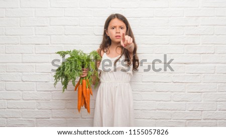 Young hispanic kid over white brick wall holding fresh carrots pointing with finger to the camera and to you, hand sign, positive and confident gesture from the front