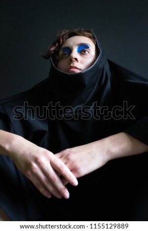 fashion photo portrait of a girl with curly hair in black clothes with blue paint in front of her eyes in a hooded hand connected
