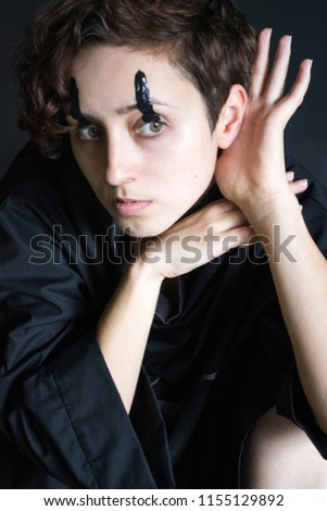 fashion photo portrait of a girl with curly short hair in black clothes with black paint over her eyes and a hand near her ear listens to a dark background