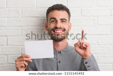 Young adult man over brick wall holding blank paper surprised with an idea or question pointing finger with happy face, number one