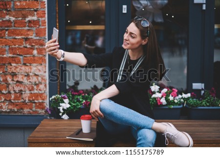 Positive female student spending time on urban setting and taking selfie pictures for own internet blog using front camera outdoors, successful hipster girl in trendy apparel photographing outdoors