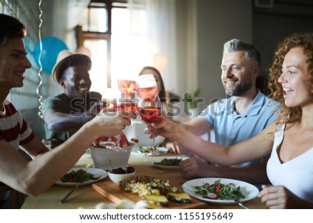 Happy young people toasting with glasses of homemade drink over served table by festive dinner