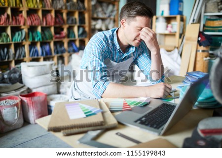Young troubled craftsman sitting in front of laptop and thinking of suitable color for one of orders