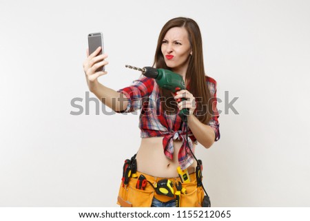 Strong excited handyman woman in shirt, kit tools belt full of instruments doing selfie on mobile phone with power electric drill isolated on white background. Female in male work. Renovation concept