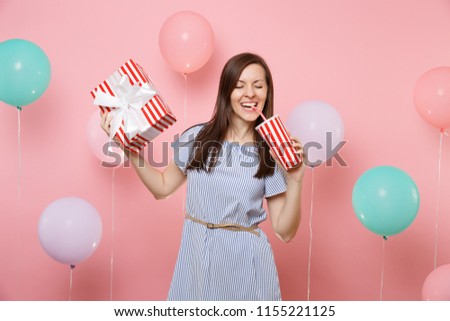 Portrait of smiling woman with closed eyes in blue dress holding red box with gift present drinking soda or cola from plastic cup on pink background with colorful air balloons. Birthday holiday party