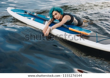 beautiful athletic tattooed young woman with blue hair relaxing on sup board in water
