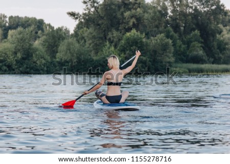 back view of blonde sportswoman sitting on sup board on river