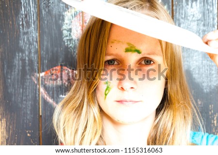 A young and beautiful girl with a face with dirty paint stands on the background of the painted paint of the wall. She covers her face from the bright rays of the sun.