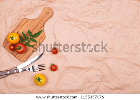 Top view of wooden cutting board (plate) with red and yellow small fresh ripe tomatoes,  knife and fork on brown crumpled paper background. Copy space.