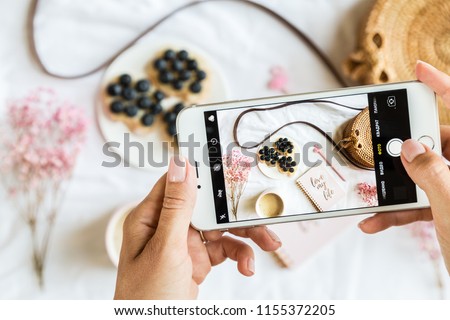Flatlay items: notebook, pen with flamingo, coffee cup, flowers pink color, wicker bag, bread on plate lying on white background, woman's hands holding mobile phone and taking picture