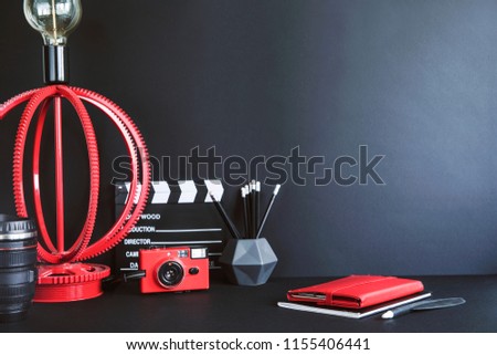 Stylish and minimalistic desk with black and red accessories, notebook, photo camera and notebooks. Black backgrounds wall. Design space in minimalistic interior.