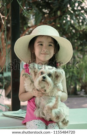 Young cute Asian girl is sitting and holding a dog with smiling. She dresses pink shirt and wears a hat. Kid and pet concept.