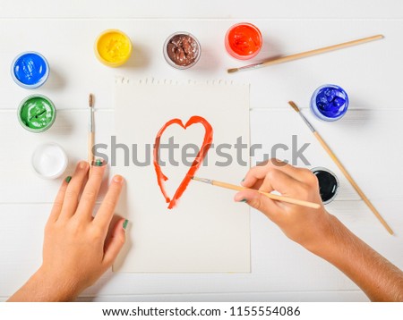 The girl draws a red outline of the heart on a white table. Flat lay.