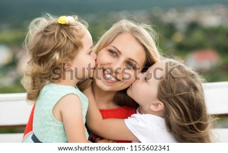 Happy childhood, family, love. Daughters kiss mother on natural landscape. Mothers day concept. Woman and girls sit on bench. Summer vacation, leisure, activity, lifestyle