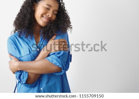 Cropped shot of stylish joyful young dark skinned woman with Afro hairstyle keeping eyes closed, hugging herself, having pleased look, posing against white blank wall with copy space for your text