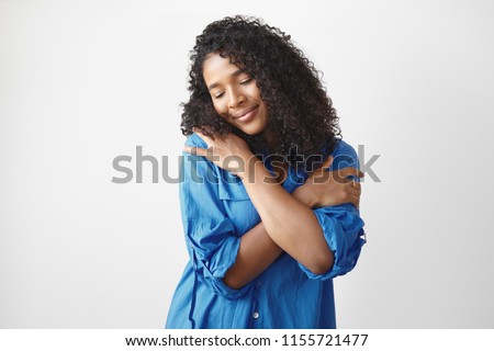 Studio shot of gorgeous charming young Afro American lady in stylish blue shirt having pleased happy facial expression, closing eyes and smiling, embracing herself, full with love and appreciation Royalty-Free Stock Photo #1155721477