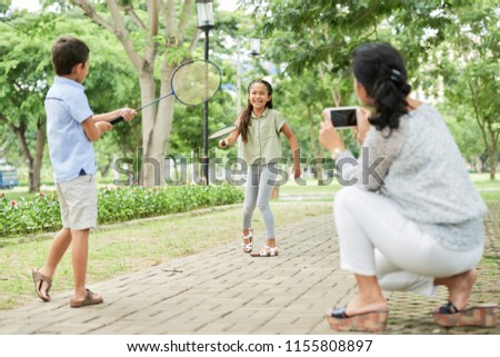 Adult woman using smartphone to take picture of cheerful Asian kids playing badminton in park