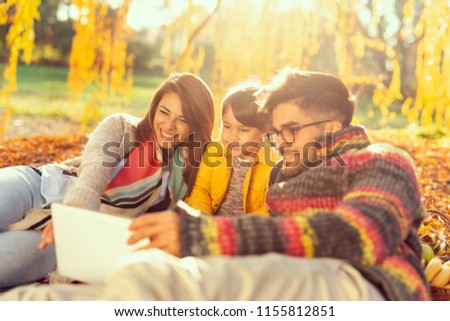 Beautiful young family lying on a picnic blanket, enjoying an autumn day in the park, watching cartoons on a tablet computer