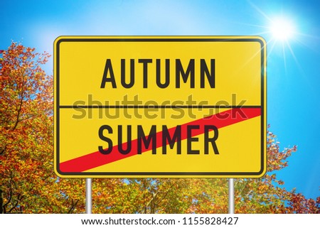 Yellow roadsign or place-name sign with autumn and summer written on it and summer being crossed out against sunny sky with colorful fall foliage in background