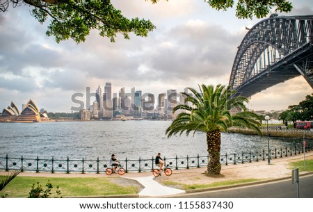 Wide Shot of Tourists Biking along Sydney Harbour, with a view of the Sydney Skyline, Milsons Point, Waterfront, Sydney Harbour Bridge, and Opera House on a Summer Evening - Sydney, Australia 