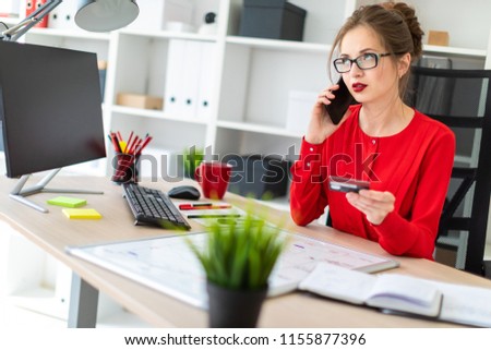 A young girl is sitting at the desk in the office, holding a bank card and phone in her hand.