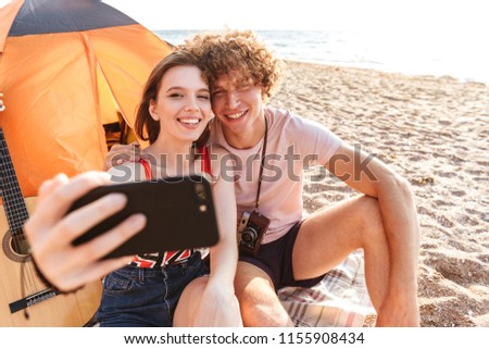 Happy young couple sitting together at the beach, camping, taking selfie