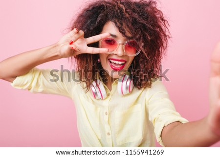 Excited black girl in cute yellow attire laughing while making selfie. Indoor portrait of gorgeous african female model taking picture of herself showing peace sign with fingers.