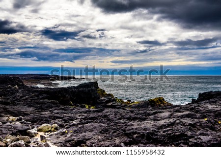 Beautiful rugged volcanic rock Iceland coastline against blue skies with low clouds. Waves pounding shoreline. Blues, greens, aquamarine colors. Lighthouse and hot steam clouds in background.